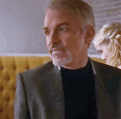 A Man In A Suit And Black Turtleneck Is Standing In Front Of A Woman . GIF
