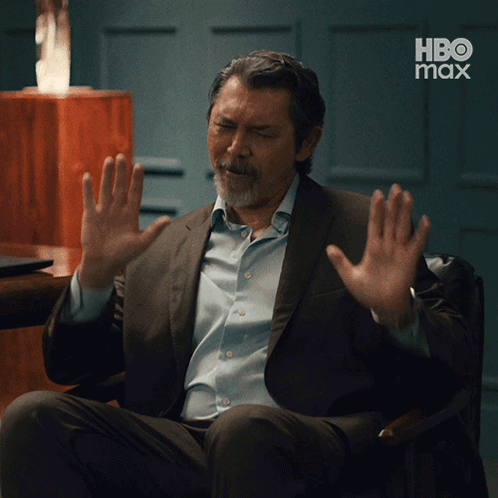 A Man In A Suit And Blue Shirt Is Sitting In A Chair With His Hands Up In Front Of A Hbo Max Logo GIF