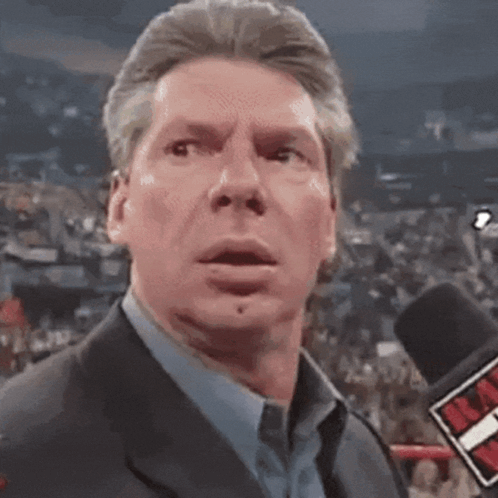 A Man In A Suit And Blue Shirt Is Talking Into A Microphone With A Red T On It GIF