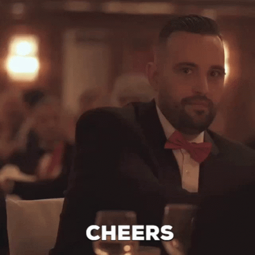 A Man In A Suit And Bow Tie Is Holding A Glass Of Wine And The Word Cheers Is Behind Him GIF
