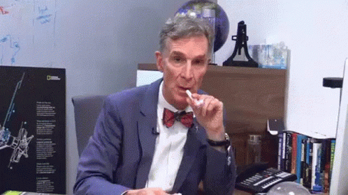 A Man In A Suit And Bow Tie Is Sitting At A Desk In Front Of A National Geographic Sign GIF