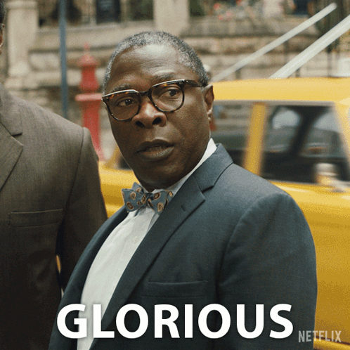 A Man In A Suit And Bow Tie Is Standing In Front Of A Yellow Car With The Word Glorious Written On It GIF