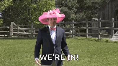 A Man In A Suit And Bow Tie Is Wearing A Pink Hat And Walking In A Field . GIF