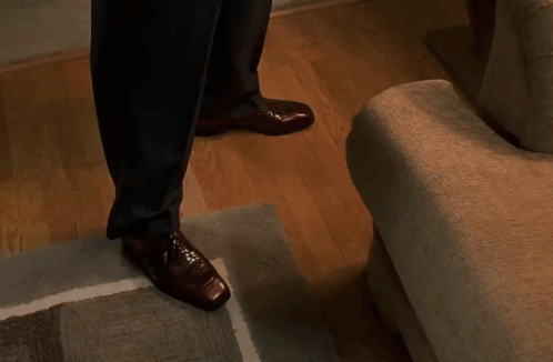 A Man In A Suit And Brown Shoes Is Walking Down Stairs In A Living Room . GIF