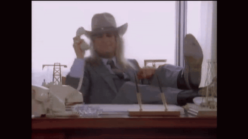 A Man In A Suit And Cowboy Hat Is Sitting At A Desk With His Feet Up Talking On A Cell Phone . GIF
