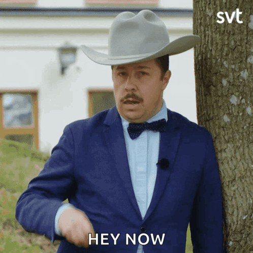 A Man In A Suit And Cowboy Hat Is Standing Next To A Tree And Says Hey Now GIF
