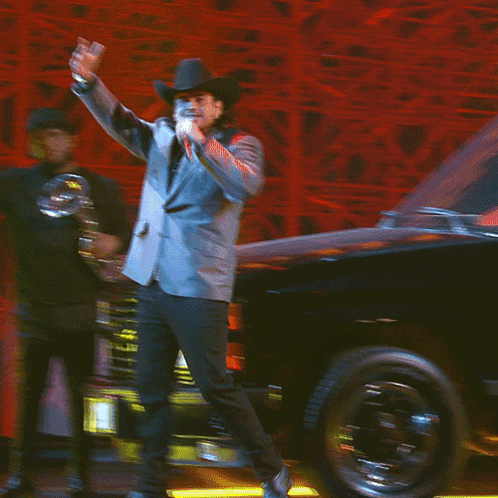 A Man In A Suit And Cowboy Hat Stands On A Stage With His Hand On His Chest GIF
