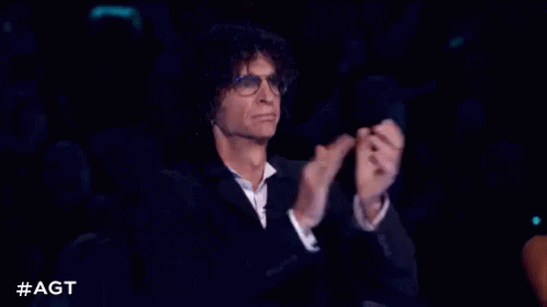 A Man In A Suit And Glasses Is Sitting In A Dark Room Clapping His Hands . GIF