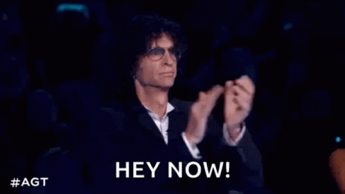A Man In A Suit And Glasses Is Sitting In A Dark Room With His Hands In The Air . GIF