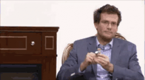 A Man In A Suit And Glasses Is Sitting In Front Of A Fireplace Holding Something In His Hand . GIF