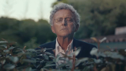 A Man In A Suit And Glasses Is Standing In Front Of A Bush With His Eyes Closed GIF