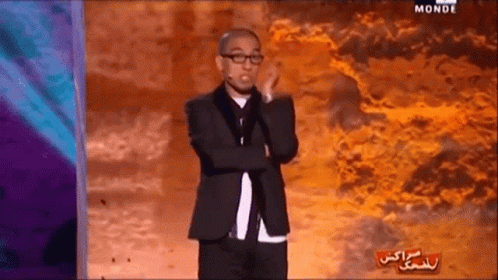 A Man In A Suit And Glasses Stands On A Stage With The Word Monde On The Bottom Left GIF