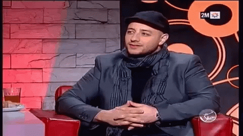 A Man In A Suit And Hat Is Sitting In A Chair With His Hands Folded In Front Of A Sign That Says 2M GIF