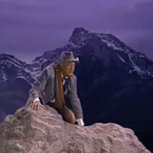 A Man In A Suit And Hat Is Sitting On Top Of A Rock . GIF