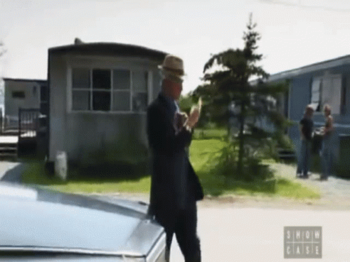 A Man In A Suit And Hat Is Standing Next To A Car With The Words Show Case On The Bottom Left GIF
