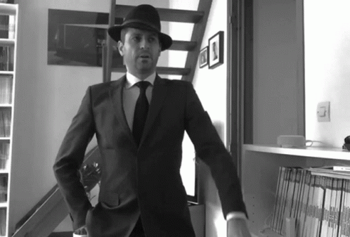 A Man In A Suit And Hat Stands In Front Of A Bookshelf With Magazines On It GIF