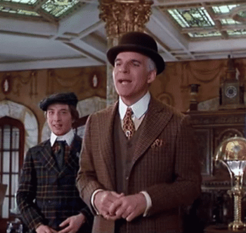 A Man In A Suit And Hat Stands Next To Another Man In A Plaid Suit GIF
