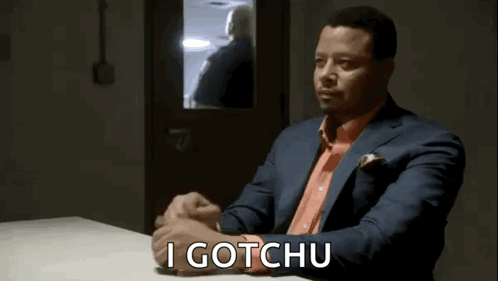 A Man In A Suit And Orange Shirt Is Sitting At A Table With The Words `` I Gotchu '' Written On It . GIF