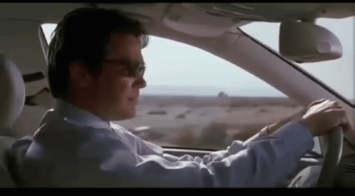 A Man In A Suit And Sunglasses Is Driving A Car On A Highway . GIF
