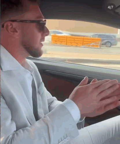 a man in a suit and sunglasses is sitting in a car with his hands folded in front of him