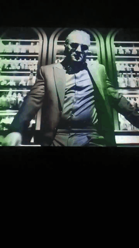 A Man In A Suit And Sunglasses Is Standing In Front Of A Display Of Bottles GIF