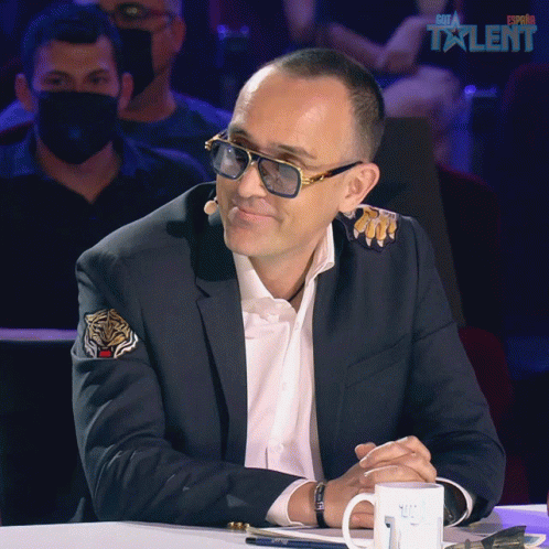A Man In A Suit And Sunglasses Sits In Front Of A Sign That Says Spain Talent On It GIF