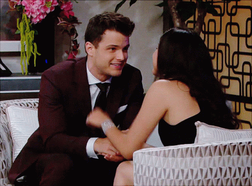 A Man In A Suit And Tie And A Woman In A Black Dress Are Sitting Next To Each Other GIF