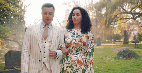 A Man In A Suit And Tie And A Woman In A Floral Dress Are Walking In A Park . GIF