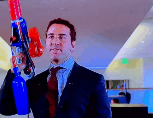 A Man In A Suit And Tie Holds A Large Toy Gun GIF