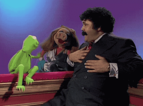 A Man In A Suit And Tie Holds His Chest While Two Muppets Look On GIF