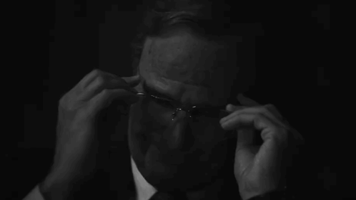 A Man In A Suit And Tie Is Adjusting His Glasses In The Dark . GIF