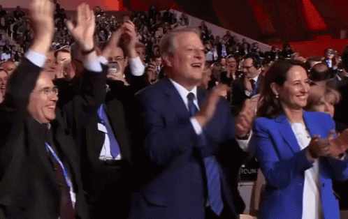 A Man In A Suit And Tie Is Applauding While A Woman In A Blue Jacket Looks On GIF