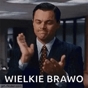 A Man In A Suit And Tie Is Clapping His Hands In Front Of A Sign That Says Wielkie Bravo . GIF