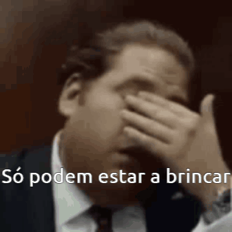 A Man In A Suit And Tie Is Covering His Face With His Hand And Says `` So Podem Estar A Brincar '' . GIF