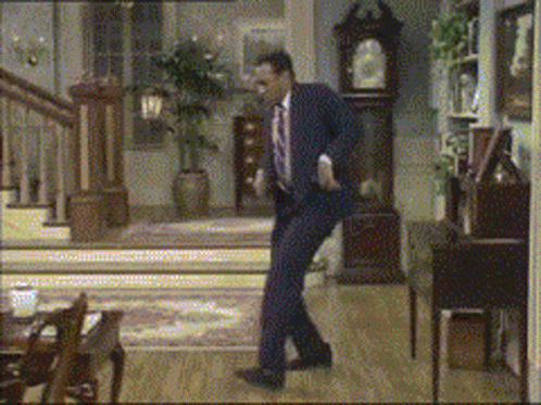 a man in a suit and tie is dancing in a living room with a clock in the background