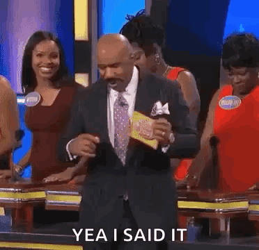 A Man In A Suit And Tie Is Dancing On A Game Show While A Group Of Women Are Standing Behind Him . GIF