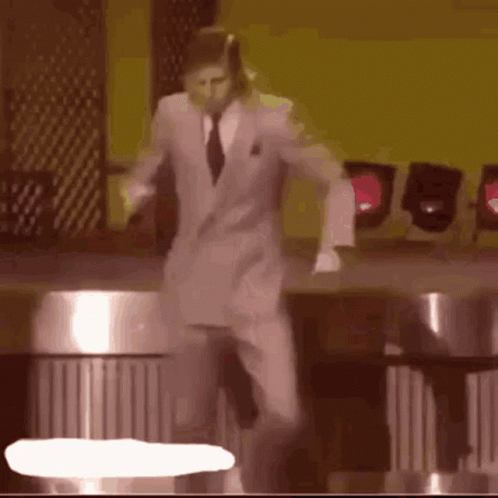 A Man In A Suit And Tie Is Dancing On A Stage In Front Of A Yellow Wall . GIF