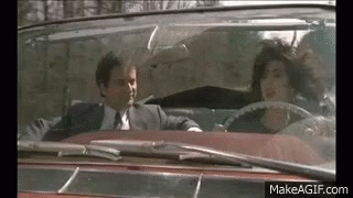 A Man In A Suit And Tie Is Driving A Convertible With A Woman In The Back Seat . GIF