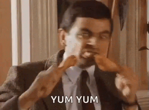 A Man In A Suit And Tie Is Eating A Chicken Leg And Saying Yum Yum . GIF