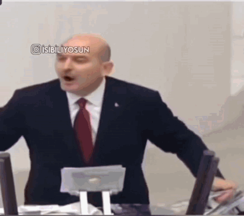 A Man In A Suit And Tie Is Giving A Speech In Front Of A Computer . GIF