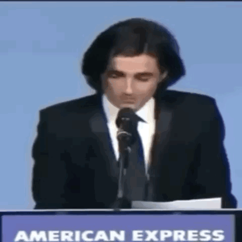 A Man In A Suit And Tie Is Giving A Speech In Front Of An American Express Sign GIF