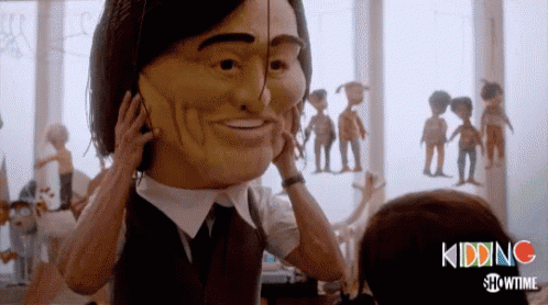 A Man In A Suit And Tie Is Holding A Large Mask On His Face GIF