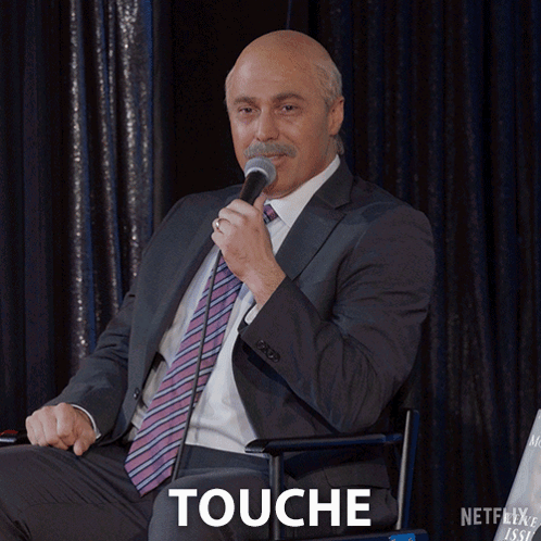 A Man In A Suit And Tie Is Holding A Microphone And The Word Touche Is Below Him GIF