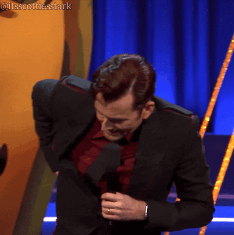 A Man In A Suit And Tie Is Holding A Microphone In Front Of A Blue Curtain That Says Itsscottiesstark On It GIF