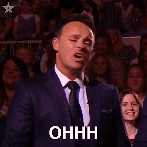 A Man In A Suit And Tie Is Holding A Microphone In Front Of A Crowd And Making A Funny Face . GIF