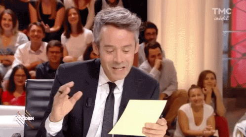 A Man In A Suit And Tie Is Holding A Piece Of Paper In Front Of A Crowd That Says Tmc On It GIF