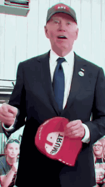 A Man In A Suit And Tie Is Holding A Red Hat That Says " I Must " GIF