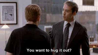 A Man In A Suit And Tie Is Hugging Another Man In A Black Shirt In An Office . GIF