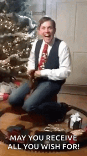 A Man In A Suit And Tie Is Kneeling In Front Of A Christmas Tree And A Train Set . GIF