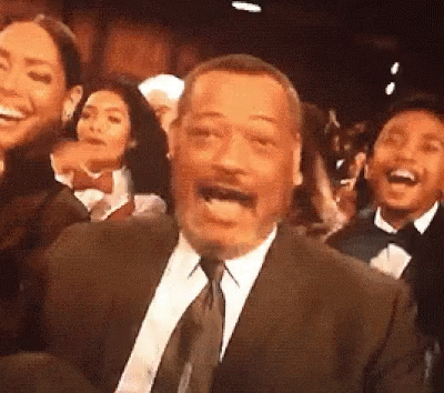 A Man In A Suit And Tie Is Laughing In Front Of A Crowd GIF